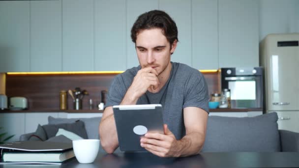 Joven europeo guapo sentado en el sofá con una tableta digital en las manos, concentrado en leer, navegar, deslizar y buscar información, descansando en casa con gadget electrónico móvil — Vídeo de stock