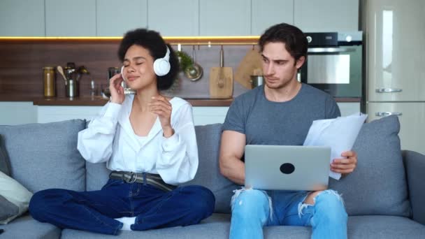Front view of freelancer guy working at laptop and graphs on paper sheets, feels bewilderment looking at his African girlfriend enjoying listening music on headphones and interrupting him from work — Stock Video