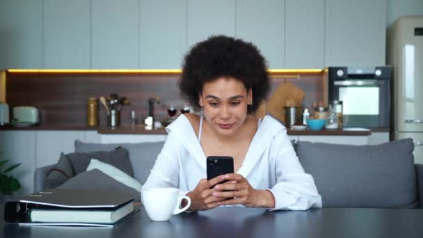 Adorable mujer afroamericana de raza mixta, escribiendo con entusiasmo texto en su teléfono inteligente, sonriendo con felicidad mientras lee grandes noticias en la pantalla, sentada en la mesa con una hermosa sonrisa en la cara — Vídeo de stock