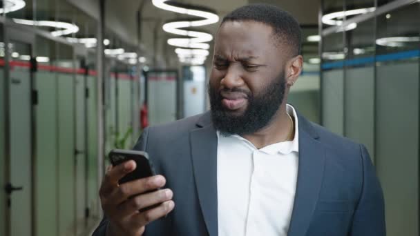 Confused African American businessman looks into a smartphone. Shocked male office worker reads an unpleasant message on a mobile phone, disgust reaction, standing in the office — Stock Video