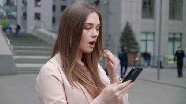 オフィスビルの外に立っているスマートフォンを見てショックを受けた若い女性。悪いニュースを持つ不快な驚きの女性. — ストック動画