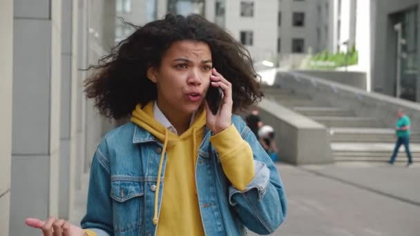 Arrabbiato giovane donna parlando su smartphone all'aperto. Irate ragazza afro-americana con i capelli ricci avendo conversazione sgradevole attraverso il telefono. Comunicazione e concetto tecnologico — Video Stock