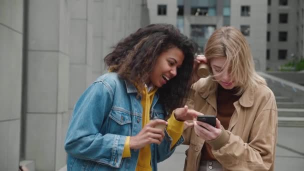 两个不同的年轻女人站在街上笑着用智能手机。两位女性朋友享受在一起的时光，看看手机的屏幕，然后讨论社交网络 — 图库视频影像