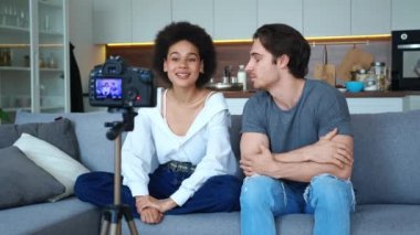 Yakışıklı erkek arkadaşıyla el sallayan güzel Afro-Amerikalı kadın tripoda monte edilmiş kameraya bakıyor oturma odasındaki kanepede birlikte oturuyor ve bir video konuşması kaydediyor.