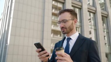 Erkek ofis çalışanı akıllı telefonuna bakıyor ve modern büyük ofis binasının yakınında kahve içiyor. Sokakta kahve ve cep telefonuyla dikilen resmi takım elbiseli iş adamının portresi..