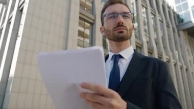 Gözlüklü ve resmi takım elbiseli kendine güvenen bir iş adamı şehir merkezinde dışarıda bekliyor. Şehir arka planında beklerken kağıt belgelerine bakan meşgul yöneticinin portresi.