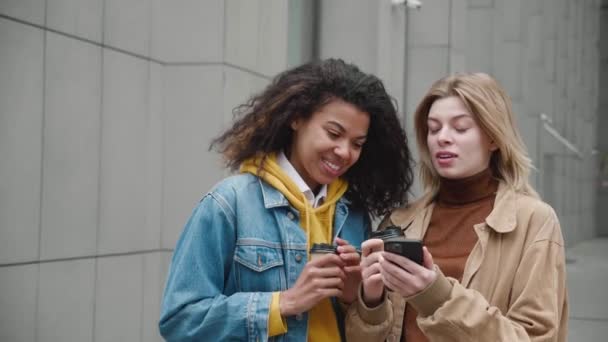 Duas jovens mulheres diversificadas sorrindo e usam o smartphone enquanto estão na rua. Duas amigas gostando de passar tempo juntas, olhar para a tela do telefone celular e discutir redes sociais — Vídeo de Stock
