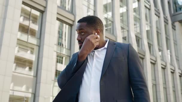 Un empresario africano seguro se pone audífonos inalámbricos para escuchar música. Retrato de hombre barbudo en traje clásico parado al aire libre en el distrito de negocios. — Vídeos de Stock