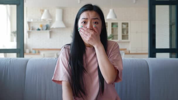 Young dark-haired with long hair woman of Asian descent looks at camera, sitting on sofa and suddenly covers her mouth being afraid and shocked by watched. Main focus on facial expression and emotion — Stock Video