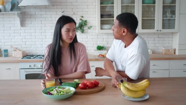 Vackra glada interracial par kär i stilig blandras afroamerikansk man och vacker asiatisk kvinna ta frukost tillsammans i ett mysigt kök, clink glas med hälsosam apelsinjuice — Stockvideo