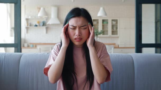 Frustrated deappointed and upset young Asian woman sitting on the couch in living room looking at camera, feeling chock, massaging her temple. Main focus on facial expressions and negative emotions — Stock Video