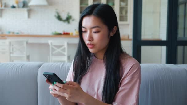 Junge nachdenkliche dunkelhaarige Asiatin in rosa T-Shirt, die auf dem Smartphone tippt, SMS verschickt und es sich zu Hause auf der Couch gemütlich macht. Schöne Frau SMS per Handy — Stockvideo