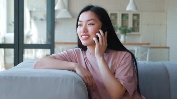 Gyönyörű és barátságos fiatal ázsiai etnikai nő rózsaszín pólóban ül a kanapén, könyökét a kanapé hátuljára támaszkodva, mobiltelefonon beszél, a többit otthon élvezi.. — Stock videók