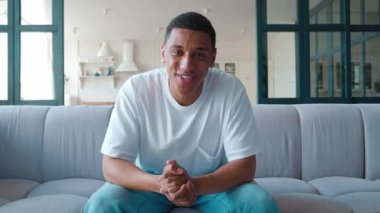 Yakışıklı ve arkadaş canlısı genç bir Afrikalı Amerikalı ya da genç bir adam evde dinleniyor, oturma odasında kanepede oturuyor ve video görüşmesinde konuşuyor, önündeki monitör kamerasına bakıyor..