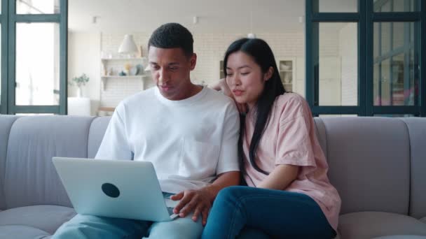 Jovem casal interracial sentado no sofá em casa com laptop e sites de surf, discutindo juntos. Jovem africano com sua namorada asiática discutindo o que eles viram na tela do monitor — Vídeo de Stock