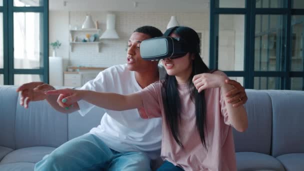 Jovem mulher asiática excitada e brincalhona vestindo óculos de realidade virtual e jogando ativamente jogos de realidade virtual, sentada ao lado de seu namorado afro-americano. Realidade Virtual e Tecnologia Digital Conceito. — Vídeo de Stock