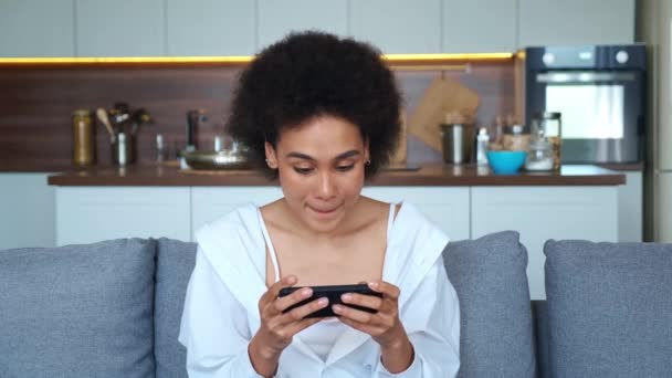 젊은 아프리카계 미국 여성 스마트폰을 하고 있는 그녀의 흥분된 모바일 애플리케이션 레이싱 게임 집 거실에서 소파에 앉아. — 비디오