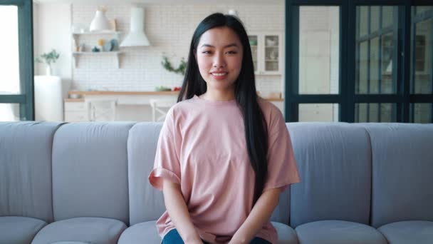 Porträt einer selbstbewussten, schönen jungen Asiatin, die zu Hause auf der Couch sitzt. Charmante junge asiatische Ethnizität Frau lächelt mit zahmem Lächeln, blickt zuversichtlich in die Kamera, sitzt auf dem Sofa zu Hause — Stockvideo