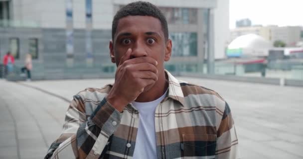 Rädd chockad ung afrikansk amerikansk man 25s bär grundläggande casual skjorta ser kameran ser något hemskt, täcker munnen med handen utomhus på stadens gata. Människor känslor livsstil koncept — Stockvideo