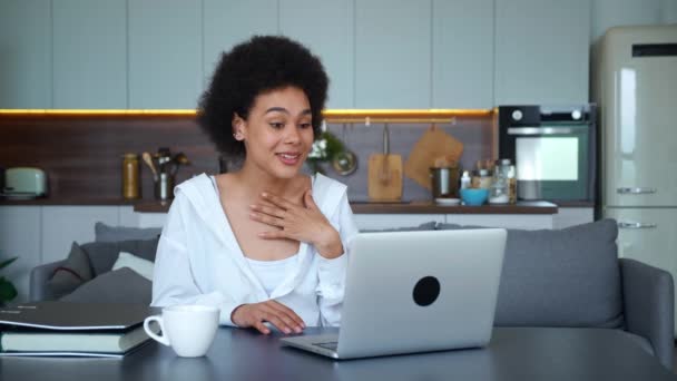 Mujer afroamericana de origen étnico sentada en la mesa de la cocina, agitando las manos mirando la cámara web usando un portátil para videollamadas. Adorable saludo de estudiante joven en línea haciendo videollamada — Vídeos de Stock