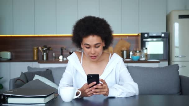 Feliz ganador de raza mixta mujer afroamericana escribiendo en el teléfono inteligente, regocijo y apretando el puño después de leer grandes noticias en la pantalla móvil, sentado en la mesa en casa con una hermosa sonrisa en la cara — Vídeo de stock
