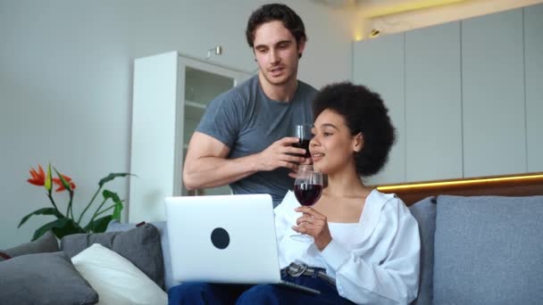 Šťastný heterosexuální pár zamilovaný do okouzlující Afričanky a pohledného Evropana, tráví spolu čas, dívají se na notebook, baví se spolu, tráví víkend doma se sklenkou vína. — Stock video