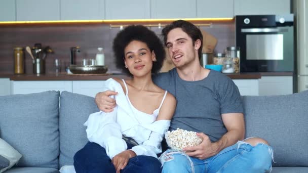 Liebenswertes heterosexuelles Paar, das sich verliebt, umarmt, auf dem Sofa sitzt, im Fernsehen über Filme spricht und Popcorn isst. Afroamerikanerin und Europäer genießen gemeinsame Zeit — Stockvideo