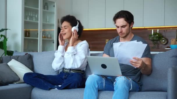 Geïrriteerde freelancer kerel werkt op laptop en grafieken op papier lakens, voelt verbijstering te kijken naar zijn Afrikaanse vriendin genieten van het luisteren naar muziek op koptelefoon en hem te onderbreken van het werk — Stockvideo