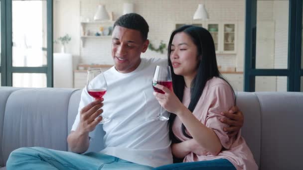 Mladé různé heterosexuální milující pár cinkat sklenice s červeným vínem na rande, objímat a sedí blízko sebe, v obývacím pokoji doma. Mladý pár má romantické chvíle spolu — Stock video