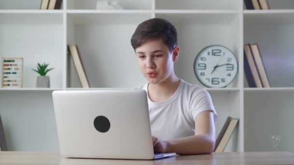 Usmívající se chlapec v chatování on-line prostřednictvím videohovoru s notebookem doma, zatímco sedí u stolu. Školák sleduje online lekce z domova. Vzdálené učení — Stock video