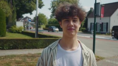 Banliyödeki parlak gündüz vakti gülümseyen genç adam portresi yeşillik ve arkası evler, doğrudan göz teması, yaklaşılabilir ifade ve rahat duruş, yaşam tarzı ve profil için ideal.