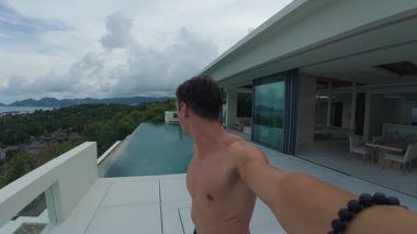Çatı katındaki sonsuzluk havuzunda selfie çeken erkek, balkon keşfi yapan vlogcu, su havuzuna dalıyor, su kabarcıkları ve sıçrama, gevşemiş tropikal atmosfer, manzara ufku.