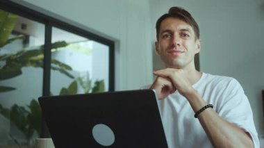 Kendine güvenen, sakince gülümseyen, modern ev ofis portresindeki bilgisayarına bakarken kameraya bakan, uzaktan kumandalı iş adamı profesyonel iş hayatı konsepti..