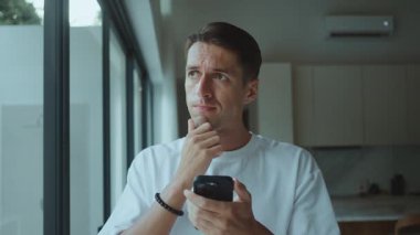 Evde akıllı telefonu kontrol eden düşünceli bir adam probleme odaklanmış hissediyor. Stres, stres kavramı endişeli erkek mesajları okuduğunu gösteriyor. Pencerenin yanında tek başına düşünen modern yaşam tarzı kaygılı.