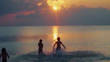 Gün batımında çocukların siluetleri yavaş çekimde denize doğru koşar. Çocuklar altın gün batımında su sıçratır. Sakin yaz akşamları. Aile tatili.