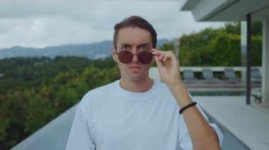 Şaşırmış bir adam güneş gözlüğü takıyor ve dışarıda modern villa ve havuzun yanında şok geçiriyor. Duygusal tepki konsepti. Şok olmuş bir adam şaşkınlık, uyarı, ani keşif anı gösterir.