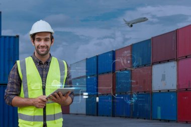 Güvenlik teçhizatında gülümseyen lojistik mühendisi, kargo konteynır sahasında üzerinde uçan uçakla akıllı nakliye yönetimi için dijital tablet kullanıyor..