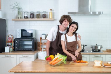 Uyumlu önlüklerle gülümseyen genç bir çift taze sebzelerle sağlıklı bir yemek hazırlıyor. Parlak, modern, beyaz bir mutfakta..