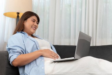 Gülümseyen hamile bir kadın evdeki bir kanepede dinlenir, bilgisayarını internetten tıbbi danışma ya da video görüşmesi için kullanırken karnına dokunur..