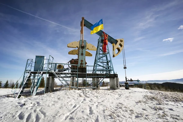 A working engineer at an oil and gas development of an old field checks the operation of mechanisms and control systems in the mountains after a winter in early spring. Flag of Ukraine yellow-blue.
