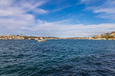 Boğazın geniş açılı manzarası, tekneler, Boğaz Köprüsü ve İstanbul kıyı şeridi parlak mavi bir gökyüzünün altında.