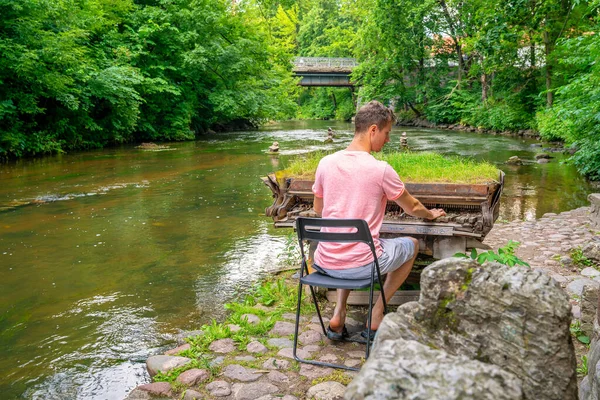 Young man playing abandoned gran piano covered with grass, rundown piano by a river bank Vilnia river in Uzupis artists quarter in Vilnius