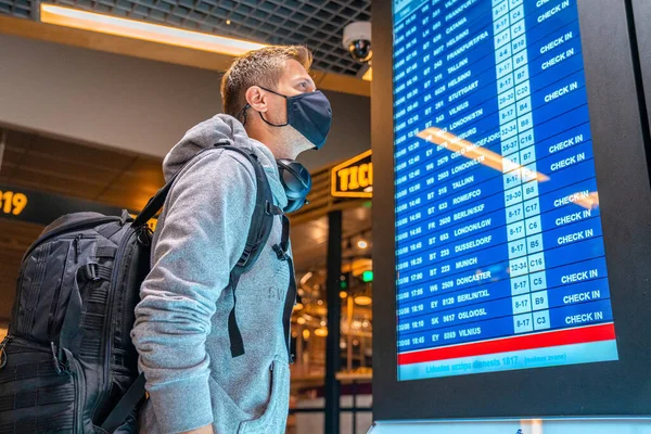 空の空港で空港情報ボード上のスーツケースチェックフライトキャンセルステータスと顔マスクを身に着けている若い男のバナー写真 航空会社の破産 航空危機または新しい通常の概念 — ストック写真