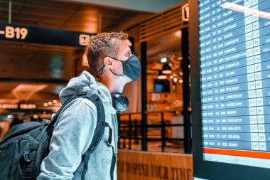 Boş havaalanındaki havaalanı bilgi panosunda yüz maskesi takan ve bavulunu kontrol eden bir gencin resmi. Uçak iflası, havayolu krizi veya yeni normal konsept