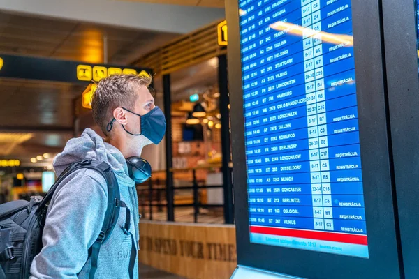 空の空港で空港情報ボード上のスーツケースチェックフライトキャンセルステータスと顔マスクを身に着けている若い男のバナー写真 航空会社の破産 航空危機または新しい通常の概念 — ストック写真