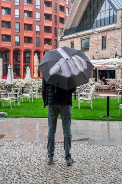 Tallinn, Estonya. 10 Haziran 2020. Öfkeli öfkeli adam şemsiyesini tutuyor ve orta parmağı havada yağmuru işaret ediyor..