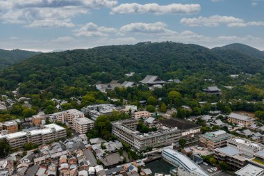 Japonya, Kyoto 'nun hava manzarası, tapınak çatıları, sık ağaçlar, modern ve geleneksel binalar, ve parçalı bulutlu gökyüzü altındaki yeşil tepeler..