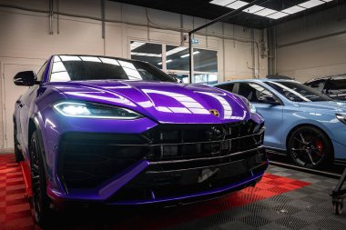 Canlı mor bir Lamborghini Urus kırmızı ve siyah kareli bir zeminde sergileniyordu. LED farlar, parlak bitiş, ve sergi salonu detayları görünür.