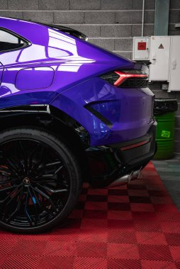 Siyah alaşımlı tekerler, çift egzoz uçları, keskin stop lambaları ve kırmızı kareli zemini olan mor Lamborghini Urus 'un arka görüntüsü..