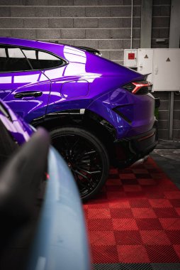 Mor Lamborghini Urus 'un arka tarafında parlak bir kaplama, kırmızı ve siyah kareli bir zemine park edilmiş. Gri tuğla duvar ve açık mavi araba görünür.